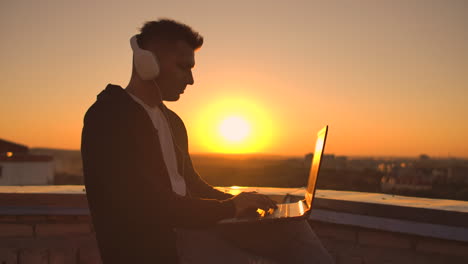 A-man-in-headphones-on-the-roof-relaxes-working-remotely-enjoying-life-despite-a-handsome-kind-of-sipping-beer-and-types-on-the-keyboard.-Trade-on-the-stock-exchange-using-a-laptop-and-enjoying-the-beautiful-view